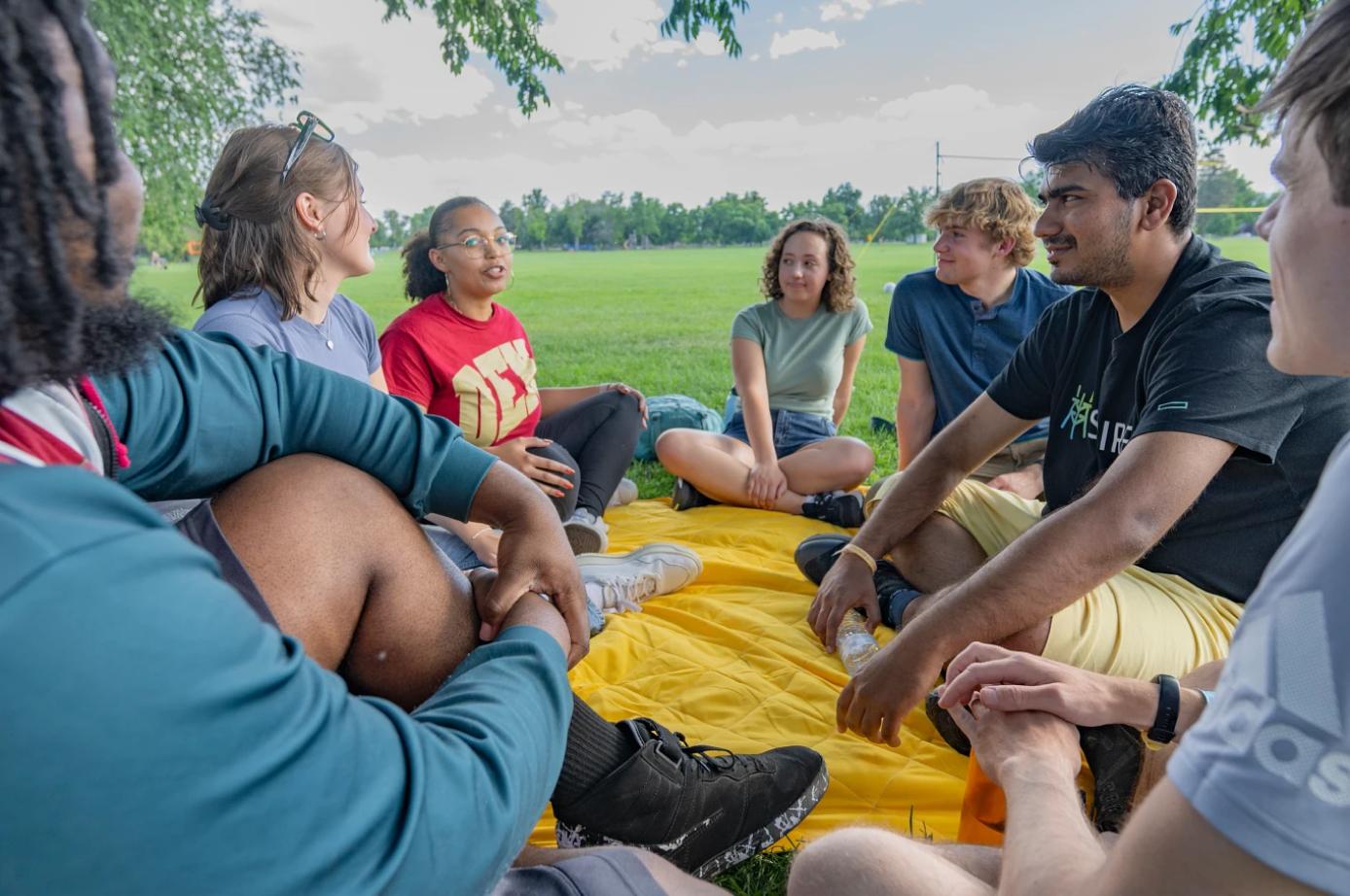 A group of students sitting in a circle on a blanket outdoors, engaging in conversation on a sunny day in a park.